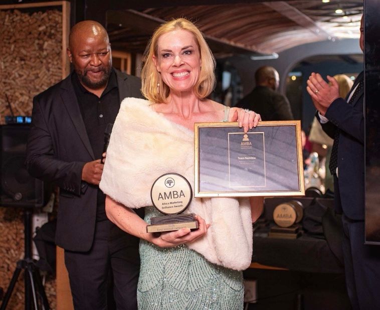 Woman in a white fur stole holds an AMBA trophy and a framed certificate at an awards ceremony, with a man in a suit behind her.