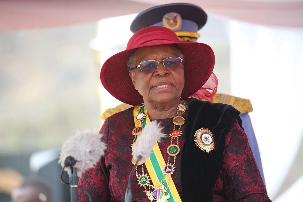 Elderly dignitary wearing a red hat and ceremonial regalia speaking at a formal event, with microphones in front.