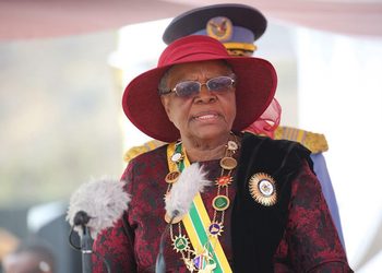 Elderly dignitary wearing a red hat and ceremonial regalia speaking at a formal event, with microphones in front.