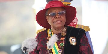 Elderly dignitary wearing a red hat and ceremonial regalia speaking at a formal event, with microphones in front.