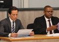 Two men in formal attire sit at a wooden table, reading papers in a formal meeting or hearing.