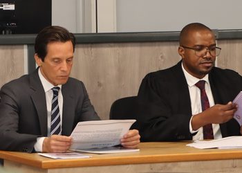 Two men in formal attire sit at a wooden table, reading papers in a formal meeting or hearing.
