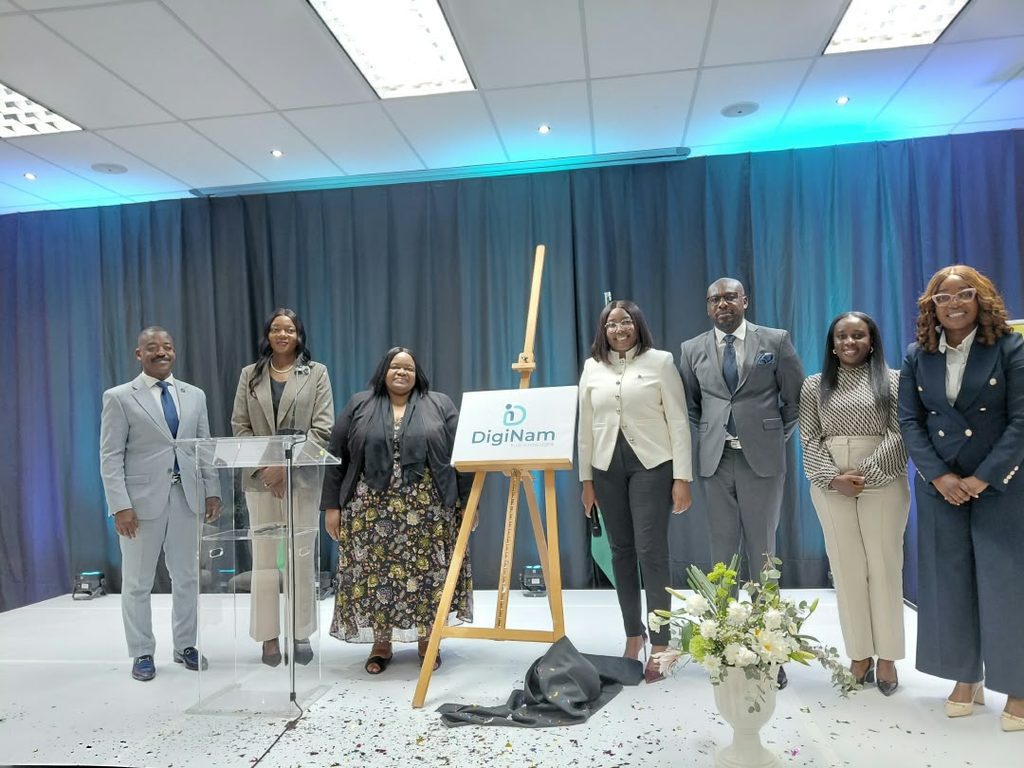 Group of eight professionals in formal attire on a stage with a DigiNam sign on an easel, unveiling a ceremony under blue curtain lighting.