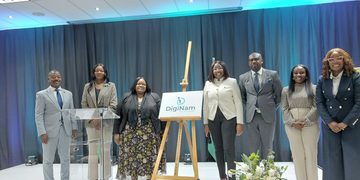 Group of eight professionals in formal attire on a stage with a DigiNam sign on an easel, unveiling a ceremony under blue curtain lighting.