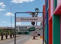Street scene with a Khomasdal sign, colorful building on the right, and a banner advertising 7 days a week under a clear blue sky.