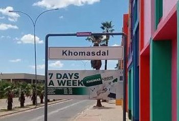 Street scene with a Khomasdal sign, colorful building on the right, and a banner advertising 7 days a week under a clear blue sky.