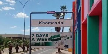 Street scene with a Khomasdal sign, colorful building on the right, and a banner advertising 7 days a week under a clear blue sky.