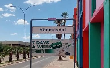 Street scene with a Khomasdal sign, colorful building on the right, and a banner advertising 7 days a week under a clear blue sky.