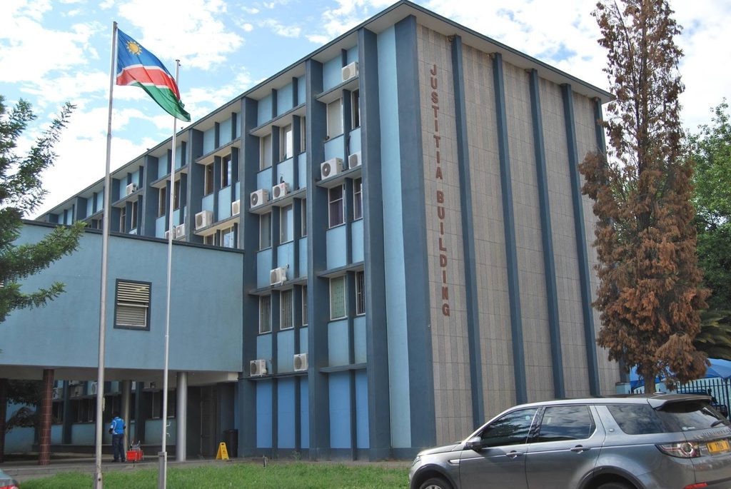 JUSTITIA BUILDING - The Brief | Namibia's Leading Business & Financial News Blue government building labeled'JUSTITIA BUILDING' with flagpoles displaying a Swaziland flag and a silver SUV in the parking area.
