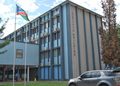 Blue government building labeled 'JUSTITIA BUILDING' with flagpoles displaying a Swaziland flag and a silver SUV in the parking area.