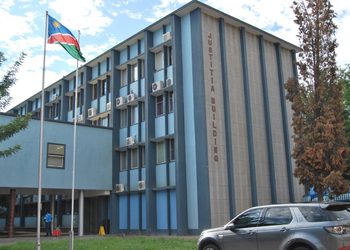 Blue government building labeled 'JUSTITIA BUILDING' with flagpoles displaying a Swaziland flag and a silver SUV in the parking area.