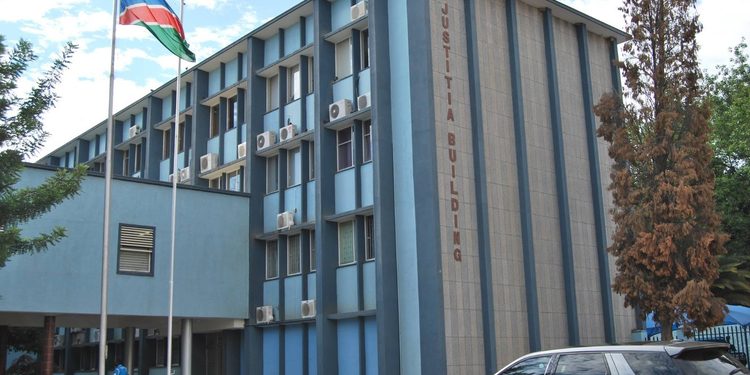 Blue government building labeled 'JUSTITIA BUILDING' with flagpoles displaying a Swaziland flag and a silver SUV in the parking area.