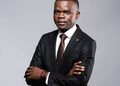 Professional portrait of a man in a dark suit, white shirt, and brown tie, standing with arms crossed and looking at the camera.
