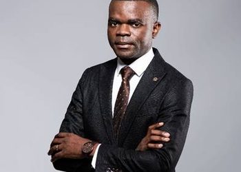 Professional portrait of a man in a dark suit, white shirt, and brown tie, standing with arms crossed and looking at the camera.