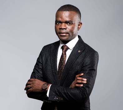 Johannes Naikaku - The Brief | Namibia's Leading Business & Financial News Professional portrait of a man in a dark suit, white shirt, and brown tie, standing with arms crossed and looking at the camera.