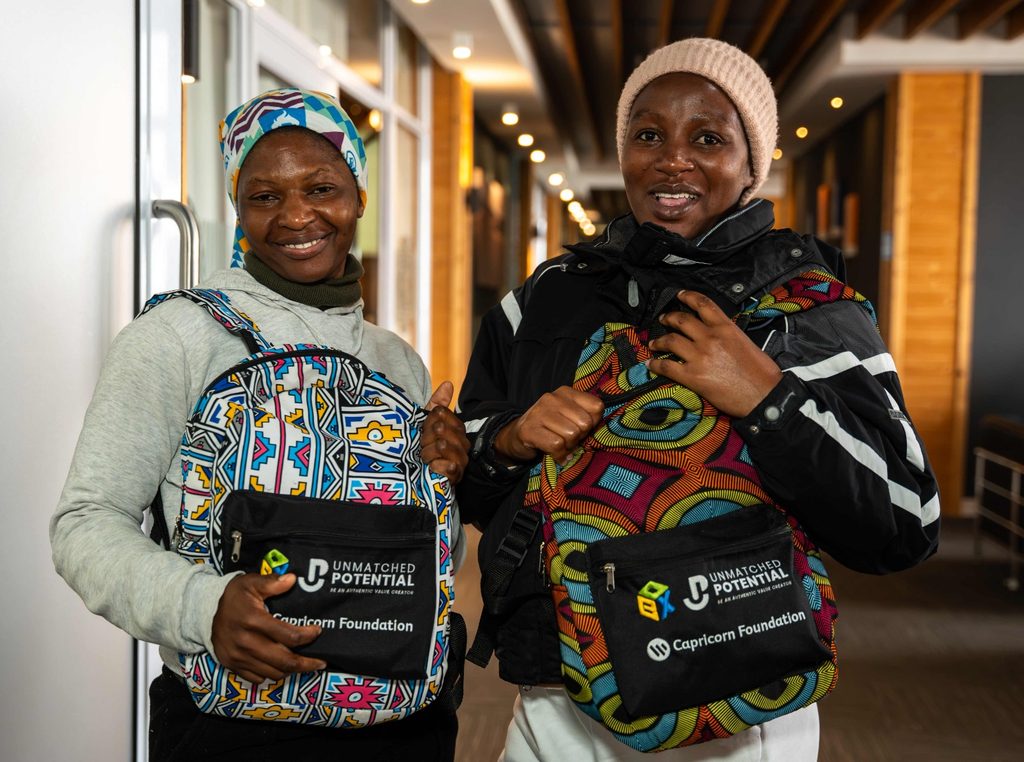 WhatsApp Image 2026-09-01 at 14734 PM - The Brief | Namibia's Leading Business & Financial News Two smiling women indoors, each wearing a colorful patterned backpack with Unmatched Potential branding and Capricorn Foundation logos visible on the pouches.