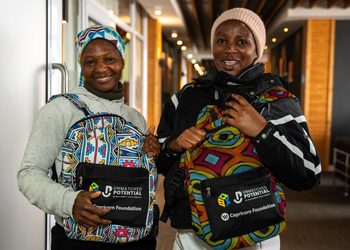 WhatsApp Image 2026-09-01 at 14734 PM - The Brief | Namibia's Leading Business & Financial News Two smiling women indoors, each wearing a colorful patterned backpack with Unmatched Potential branding and Capricorn Foundation logos visible on the pouches.