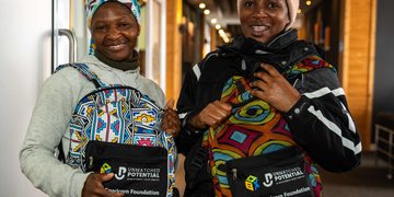Two smiling women indoors, each wearing a colorful patterned backpack with Unmatched Potential branding and Capricorn Foundation logos visible on the pouches.