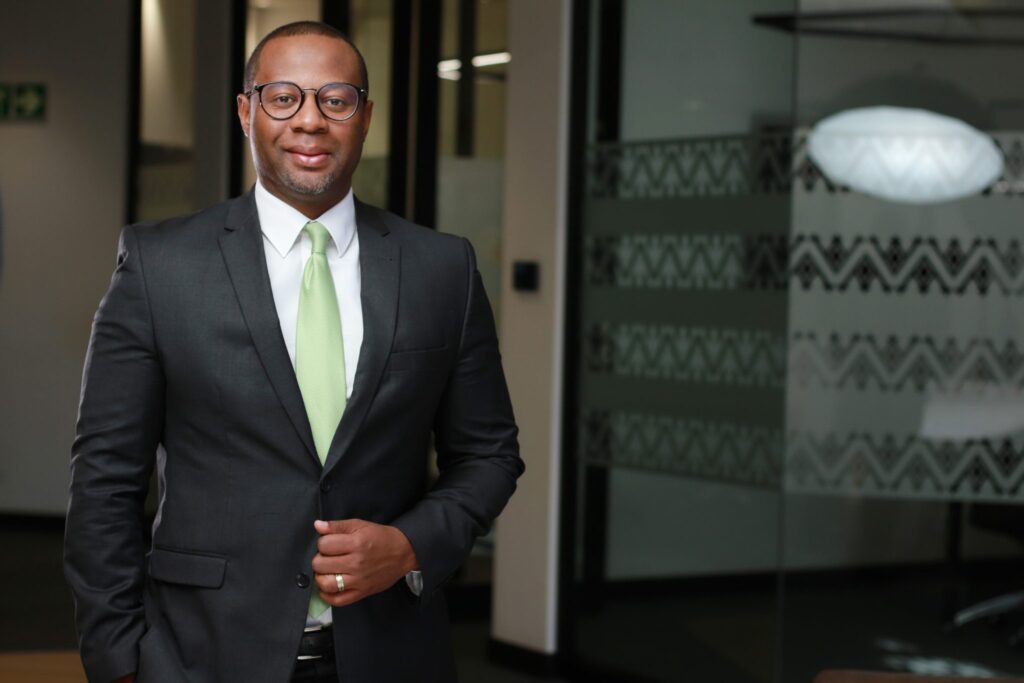 SAM IKELA - The Brief | Namibia's Leading Business & Financial News A man in a dark suit and light green tie stands in a modern office lobby, smiling softly at the camera.