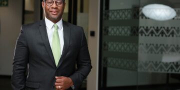 A man in a dark suit and light green tie stands in a modern office lobby, smiling softly at the camera.