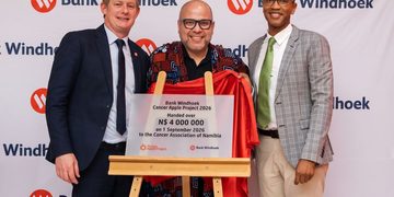 Three men pose with a draped plaque on a wooden easel at a Bank Windhoek event, the red cloth pulled aside to reveal the donation plaque.