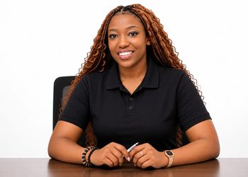 Professional portrait of a smiling woman with braided hair, seated at a desk in a black polo shirt.