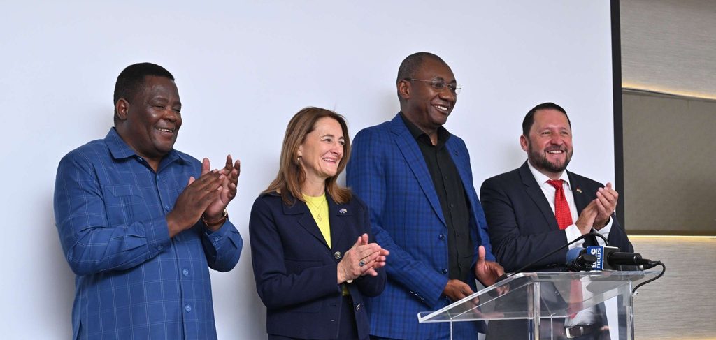 Four people stand at a podium, smiling and clapping after a presentation or press conference.