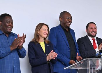 Four people stand at a podium, smiling and clapping after a presentation or press conference.