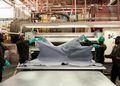 Workers in hard hats and gloves stretch a large gray fabric over a worktable in an industrial textile setting.
