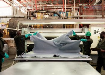 Workers in hard hats and gloves stretch a large gray fabric over a worktable in an industrial textile setting.