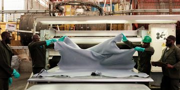 Workers in hard hats and gloves stretch a large gray fabric over a worktable in an industrial textile setting.