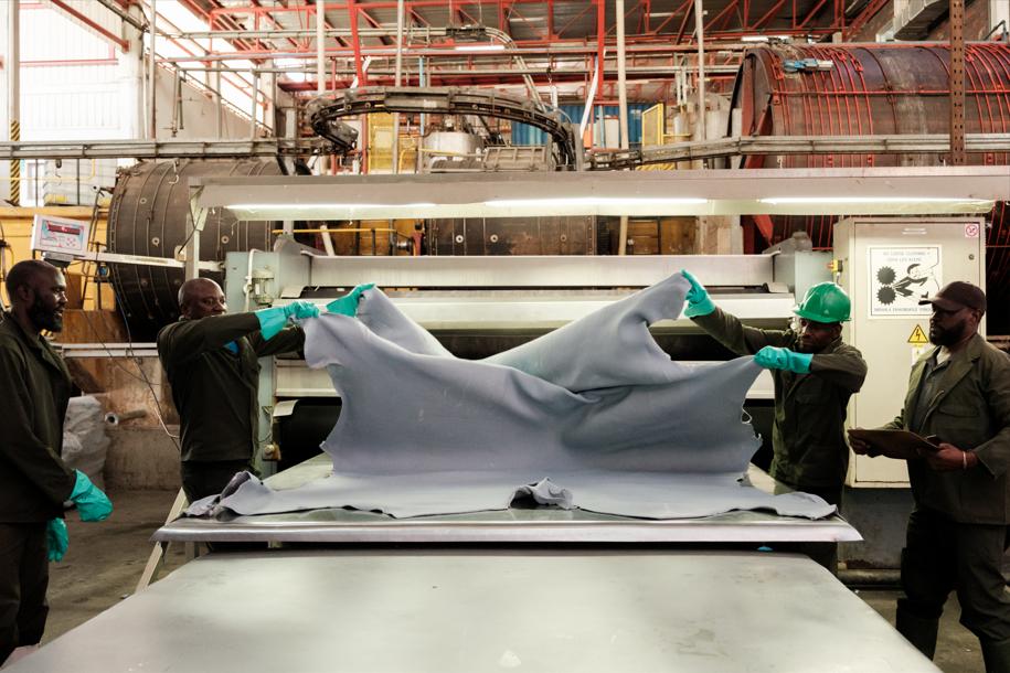 Workers in hard hats and gloves stretch a large gray fabric over a worktable in an industrial textile setting.