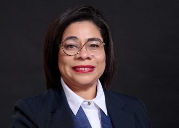 Professional portrait of a woman with short dark hair and glasses, wearing a navy blazer and white shirt against a dark backdrop.