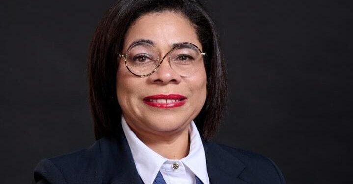 Professional portrait of a woman with short dark hair and glasses, wearing a navy blazer and white shirt against a dark backdrop.