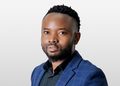 Oswald Siku Mughongora - The Brief | Namibia's Leading Business & Financial News Professional headshot of a man with short curls and beard wearing a navy blazer and black shirt against a light gray backdrop.