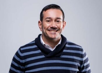 Smiling man with short dark hair wearing a navy and gray striped sweater, light background.