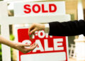 Two people exchange house keys in front of a 'Sold' sign, signaling a completed real estate sale or handover.