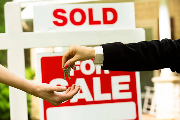HOME SALE - The Brief | Namibia's Leading Business & Financial News Two people exchange house keys in front of a 'Sold' sign, signaling a completed real estate sale or handover.