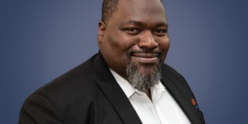 Professional headshot of a smiling Black man in a black blazer and white shirt against a blue gradient background.