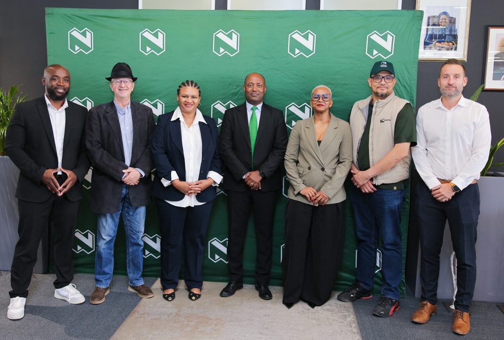 Seven diverse professionals pose in a row in front of a green backdrop with repeating white logo marks.