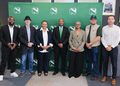 Seven diverse professionals pose in a row in front of a green backdrop with repeating white logo marks.