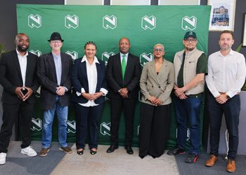 Seven diverse professionals pose in a row in front of a green backdrop with repeating white logo marks.