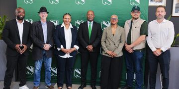 Seven diverse professionals pose in a row in front of a green backdrop with repeating white logo marks.
