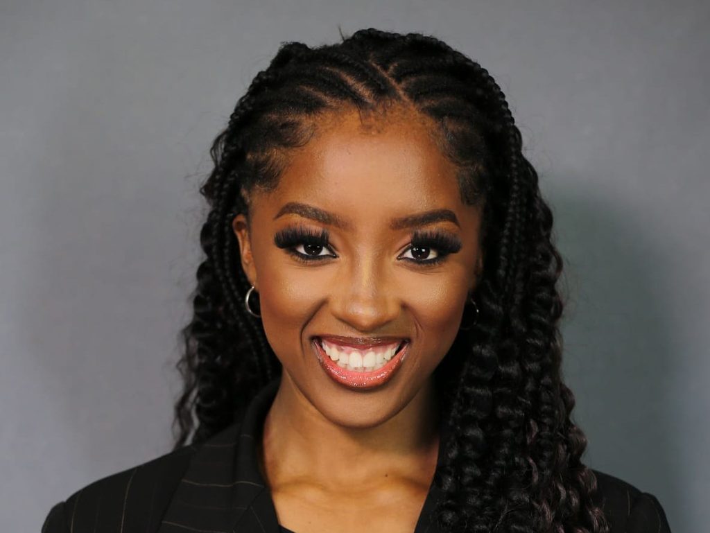 Pecky Nghaamwa - The Brief | Namibia's Leading Business & Financial News Smiling Black woman with long braids and hoop earrings, wearing a dark pinstripe blazer against a gray backdrop.