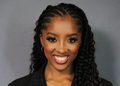 Smiling Black woman with long braids and hoop earrings, wearing a dark pinstripe blazer against a gray backdrop.