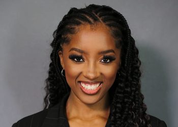Pecky Nghaamwa - The Brief | Namibia's Leading Business & Financial News Smiling Black woman with long braids and hoop earrings, wearing a dark pinstripe blazer against a gray backdrop.