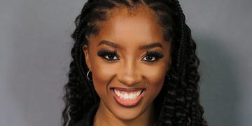 Smiling Black woman with long braids and hoop earrings, wearing a dark pinstripe blazer against a gray backdrop.