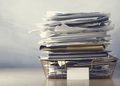 Tall stack of scattered papers and folders in a metal filing basket on a desk.