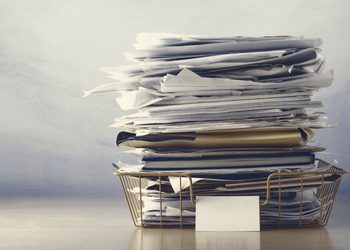 PAPERS - The Brief | Namibia's Leading Business & Financial News Tall stack of scattered papers and folders in a metal filing basket on a desk.