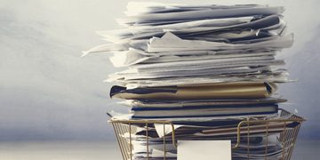 Tall stack of scattered papers and folders in a metal filing basket on a desk.
